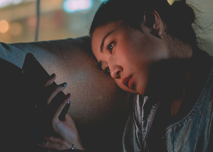 Woman lying on a couch at night, looking thoughtfully at her phone, symbolizing a messy love triangle situation.