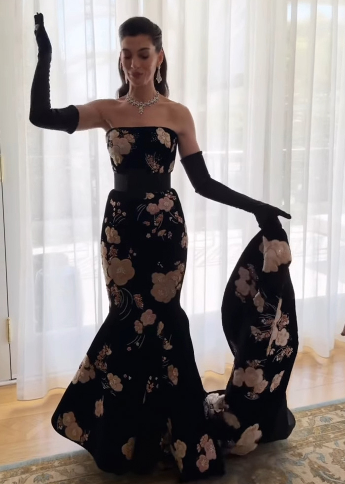 Anne Hathaway usando um vestido preto floral e luvas longas, posando dentro de casa revelando truques por trás do rosto tenso.
