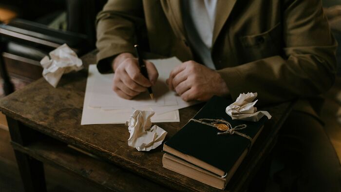 Person writing on paper at a wooden desk with crumpled sheets and a black book, illustrating unwanted.