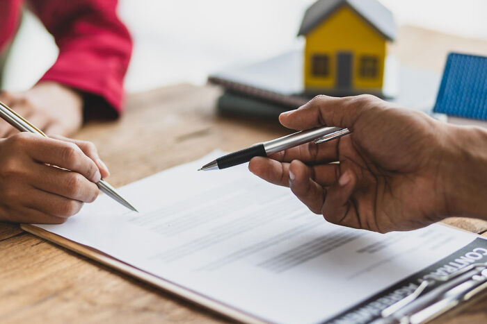 Two people holding pens over a contract on a wooden table, illustrating sabotage and karma concepts.
