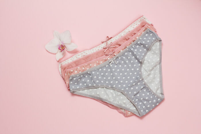 Stack of women's period underwear in pastel colors with heart patterns on a pink background and a white orchid flower.