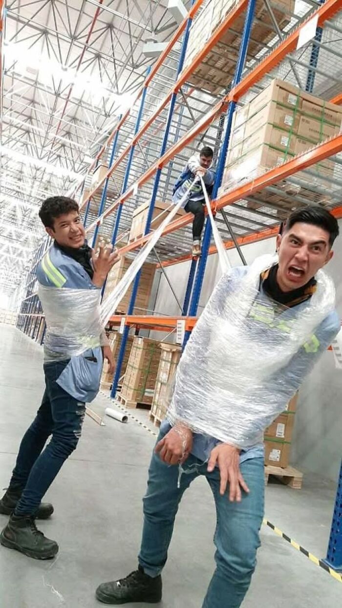 Three men in a warehouse creatively wrapped in plastic wrap, pulling a hilarious and creative pose for a fun photo.
