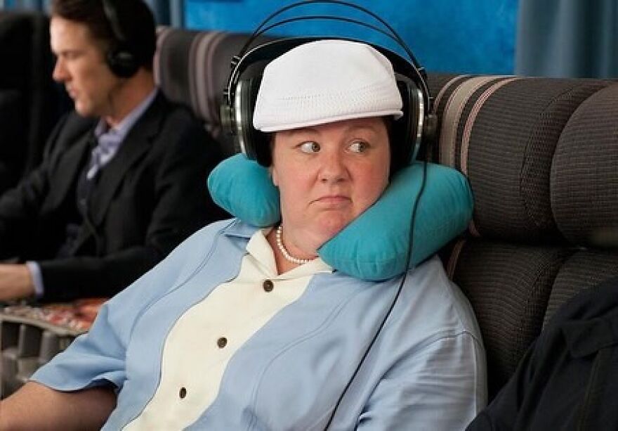 Supporting actor wearing headphones and a white cap, sitting on a plane with a travel pillow in a scene highlighting supporting performances.