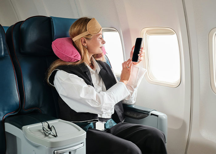 Passenger using smartphone by airplane window with neck pillow and eye mask, reflecting a major airline new rule onboard.