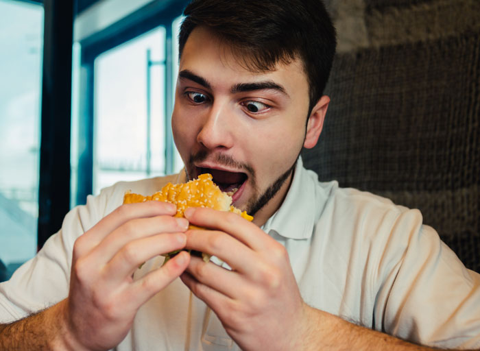 Young man eating a burger with wide eyes, illustrating weird and mildly concerning things relationships taught women about men.