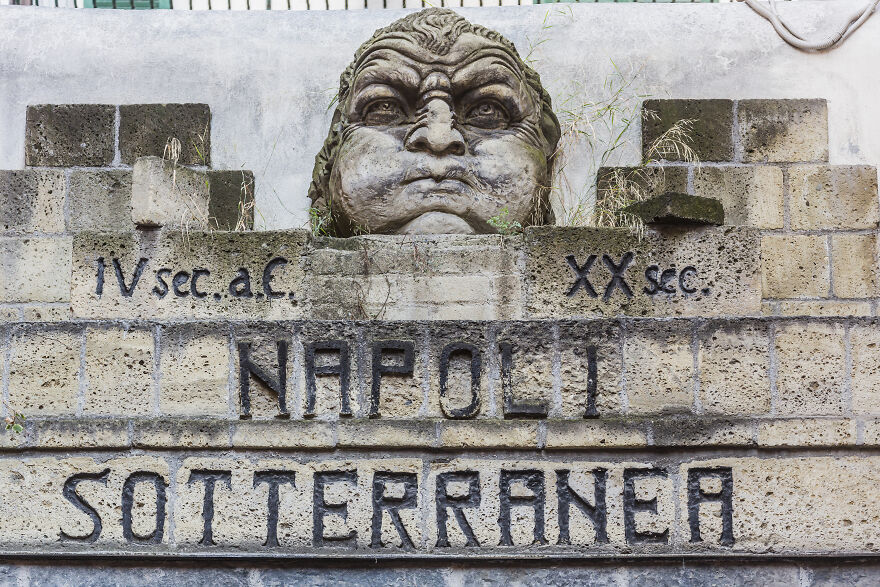 Stone sculpture and ancient stone wall with Napoli Sotterranea sign highlighting underground cities history.