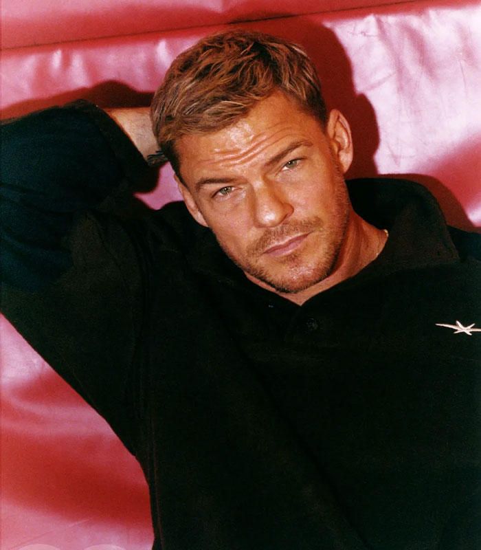 Actor Alan Ritchson reclining on a red couch, looking directly at the camera in a casual black sweater.