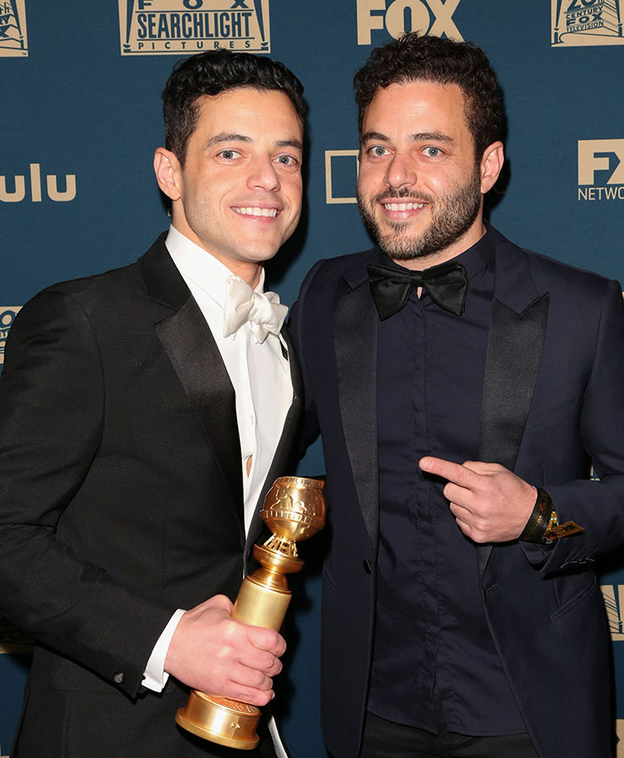 Two stars dressed in tuxedos posing together, one holding an award, highlighting twin stars the world might be stunned to find.