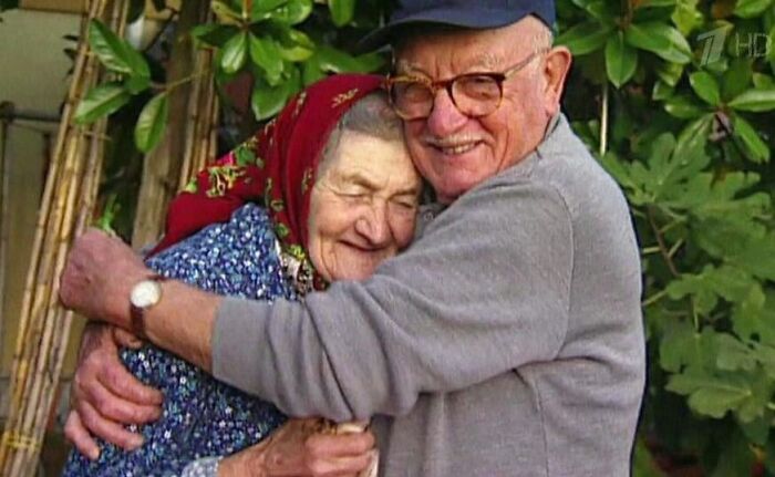 Elderly couple sharing a warm embrace outdoors surrounded by green leaves, showing deep acts of love and affection.