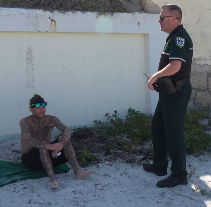 Man accused of horrific crimes sitting on Daytona Beach sand talking to a sheriff in uniform during daytime.