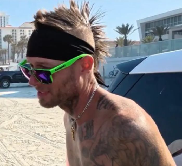 Man with mohawk hairstyle and tattoos partying with spring breakers at Daytona Beach during a lively beach event.