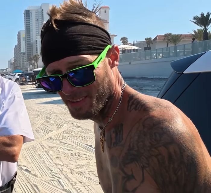 Man with tattoos wearing green sunglasses and black headband partying with spring breakers at Daytona Beach on a sunny day