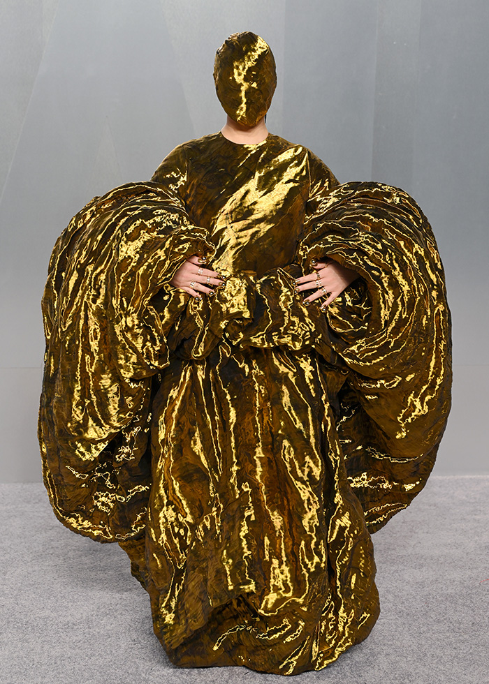 Person wearing a gold metallic outfit with exaggerated sleeves and face covering, showcasing fashion fails from Oscars afterparties.