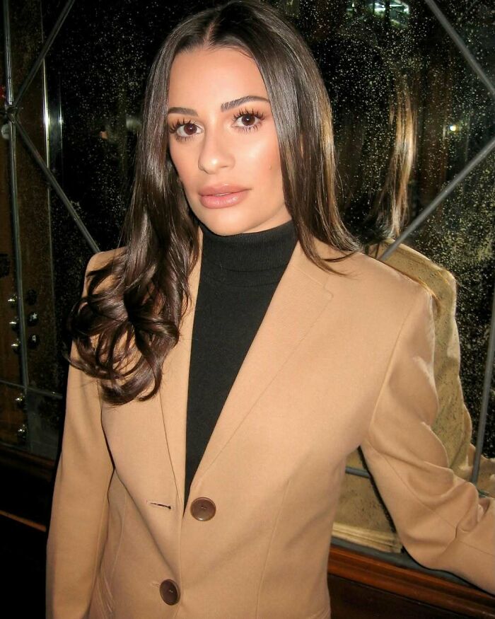 Woman in a beige blazer and black turtleneck posing indoors with reflective glass panels behind her, luxury style.