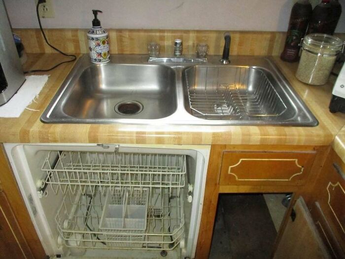 Kitchen sink above dishwasher with empty racks and outdated wooden countertop, illustrating home design disasters and poor aesthetics.