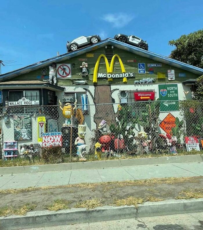 House with chaotic home design disasters featuring random signs, toys, and McDonald's logo on the roof under a clear sky.