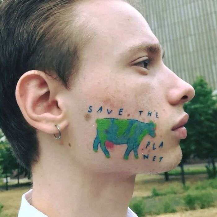 Young man with a blue and green cow tattoo on his cheek featuring a misspelled environmental message ink fail.