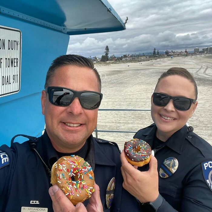 Two American police officers smiling on a beach holding colorful sprinkled donuts, showing real American things from movies.