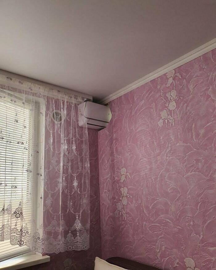 Air conditioner poorly installed in corner of pink room with floral wallpaper and lace curtains, home design disasters.