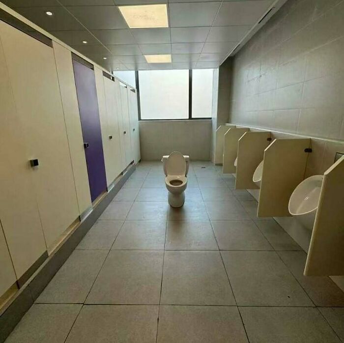 Toilet placed oddly in the middle of a public restroom with urinals and stalls, a cursed photo that makes zero sense.
