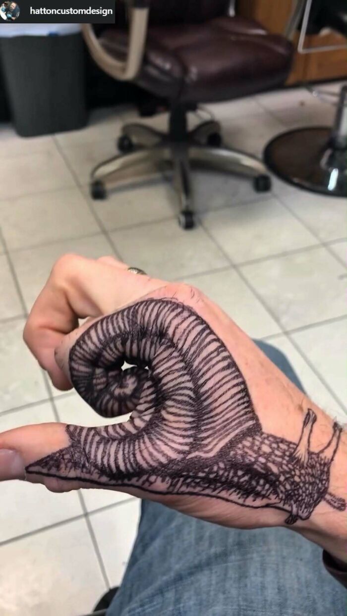 Hand with a poorly done ink tattoo of a snail twisting around the thumb in a tattoo shop setting.