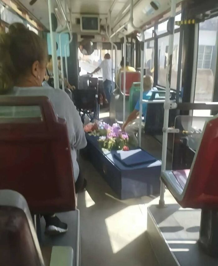 On a bus interior, a coffin with flowers creates a cursed photo moment that makes absolutely zero sense.