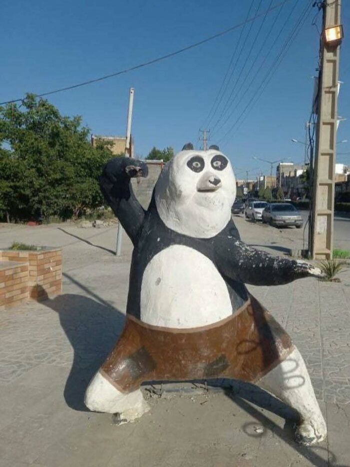 Cursed photo of a poorly sculpted panda statue in a dynamic pose on a sunny street corner.