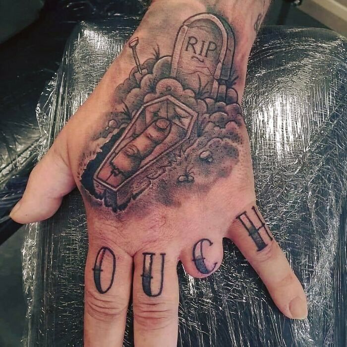 Hand tattoo with a coffin, tombstone reading RIP, and fingers spelling OUCH, showcasing ink fails and permanent regrets.