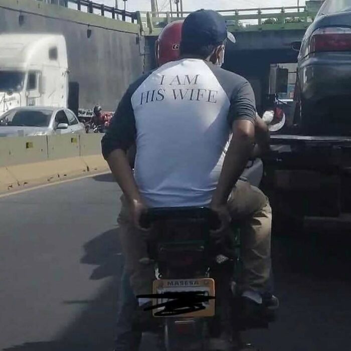Man wearing a shirt saying I am his wife riding a motorcycle in traffic, one of the cursed photos that make zero sense.
