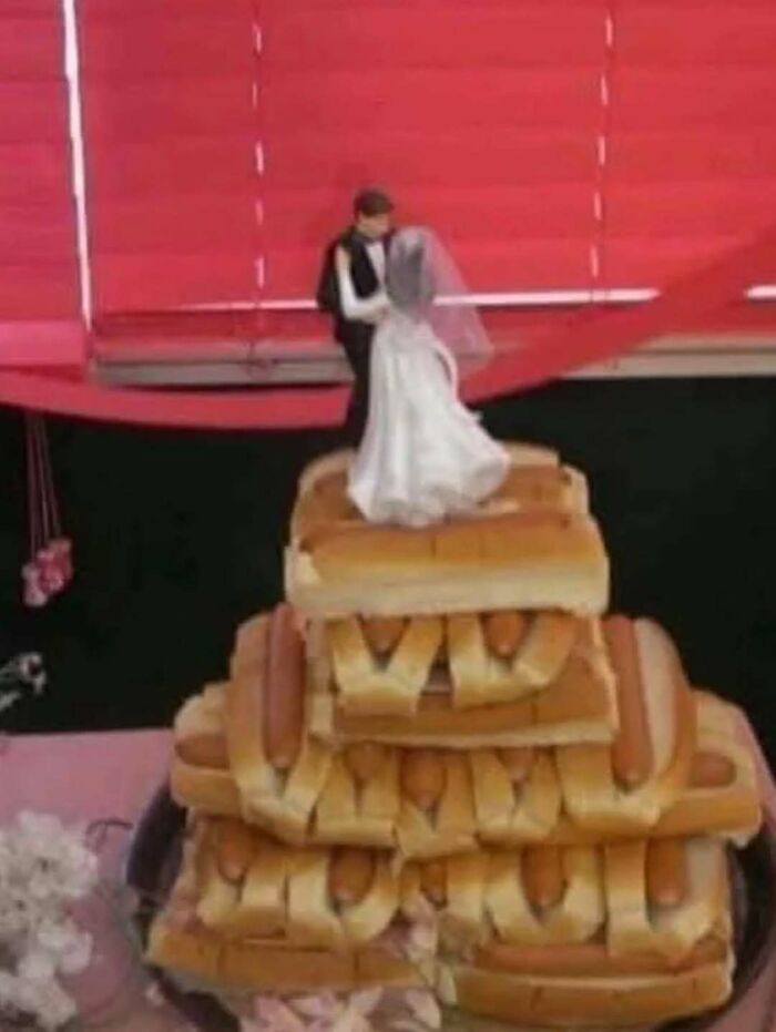 Bride and groom figurines standing on a large stack of hot dogs, creating a cursed photo with confusing perspective.
