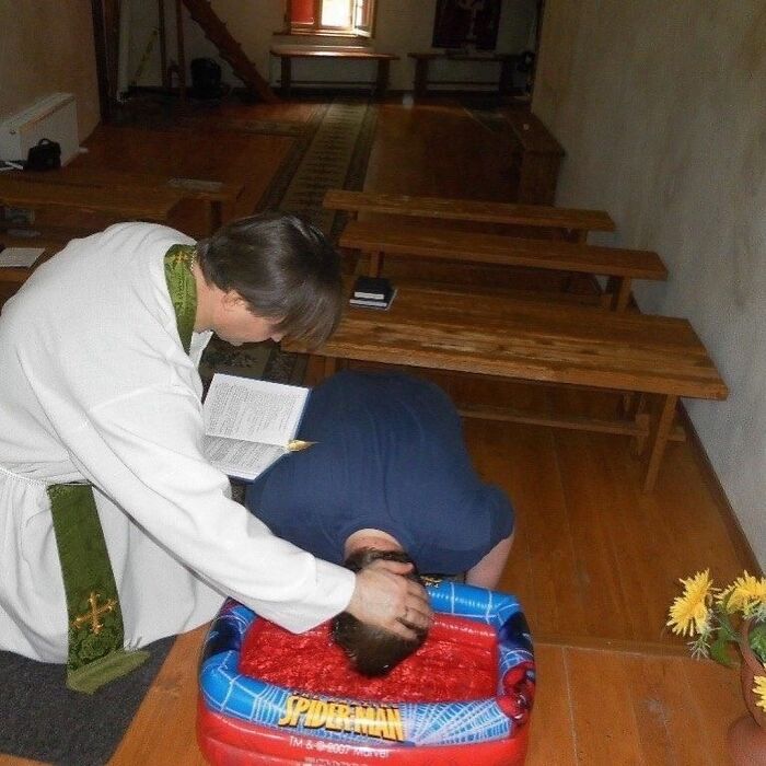 Man being baptized face-first in a small Spiderman kiddie pool in a setting that looks unusual and cursed.