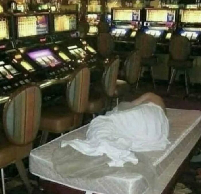 Person sleeping on a mattress in a casino surrounded by slot machines in a cursed photo that makes zero sense.