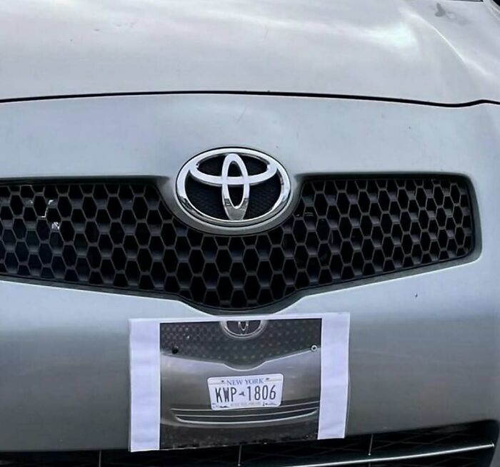 Close-up of a car with a printed paper of its own license plate taped below the actual plate in a cursed photos moment.
