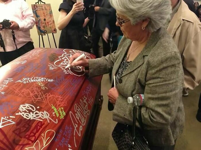 Woman signing a graffiti-covered coffin surrounded by people, a bizarre cursed photo that makes zero sense.