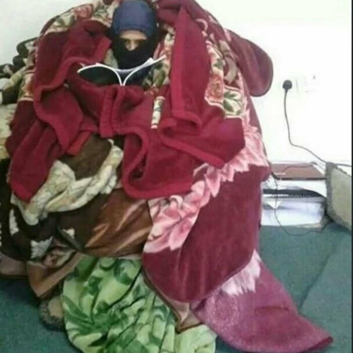 Person wrapped in multiple colorful blankets sitting on the floor with only their face visible in a cursed photo.