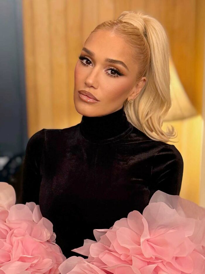 Blonde woman with high ponytail wearing black velvet top and holding large pink fabric flowers indoors.