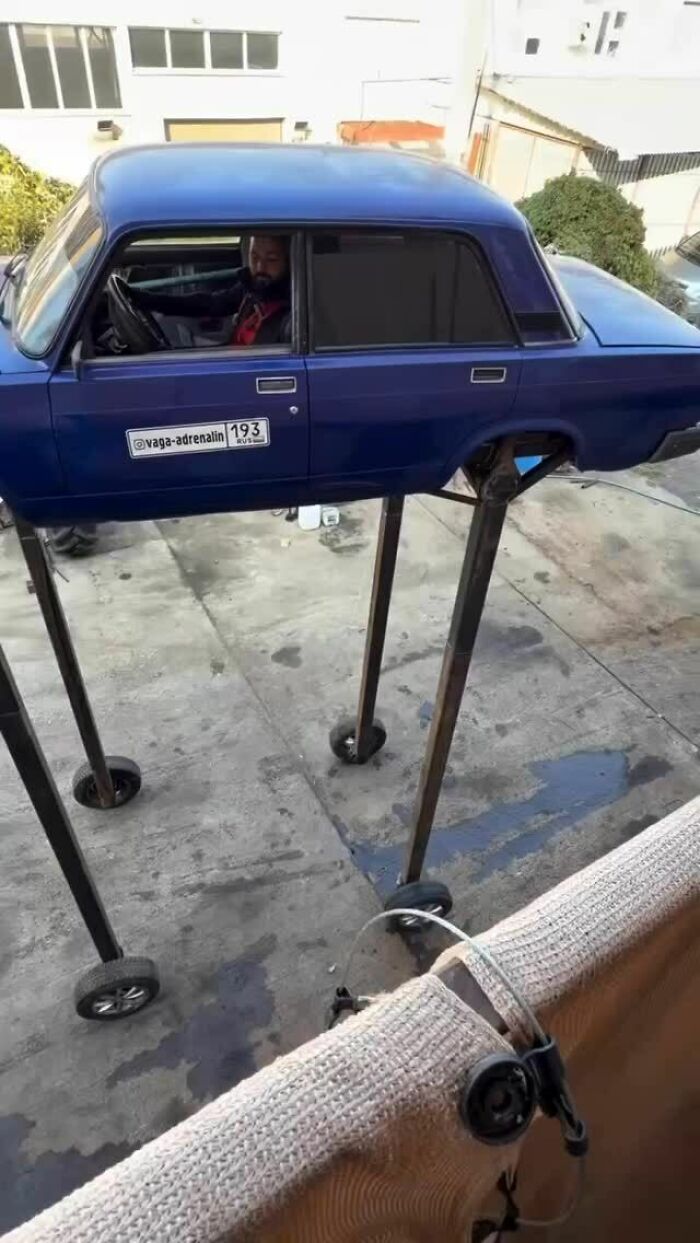 Blue car body elevated on tall metal legs with wheels, a man sitting inside, a cursed photo puzzling the viewer.