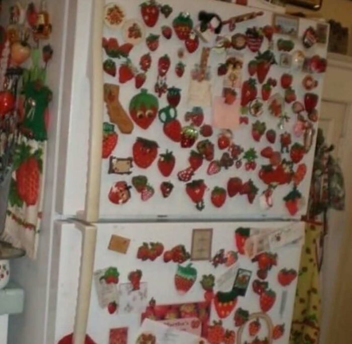 Refrigerator covered entirely with various strawberry magnets and decorations in a cluttered kitchen setting.
