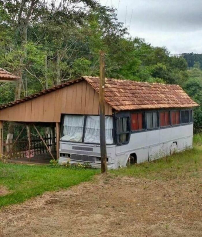 Bus converted into a house with a tiled roof and curtains, surrounded by trees and grass in a rural setting.