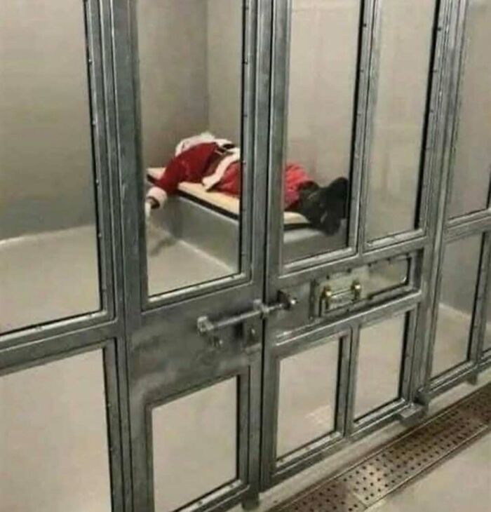 Person dressed as Santa Claus lying face down on a bench in a locked jail cell, a cursed photo that confuses viewers.