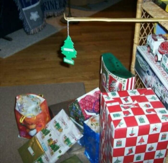 Cursed photo showing a Christmas tree air freshener hanging from a clothes drying rack above wrapped gifts on a carpeted floor.