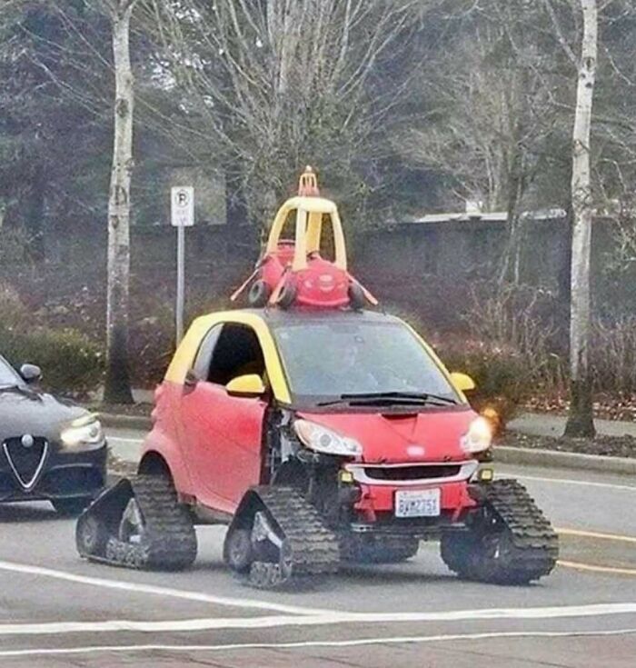 Small car with tank tracks and toy car on top driving on a street, a cursed photo that makes zero sense.