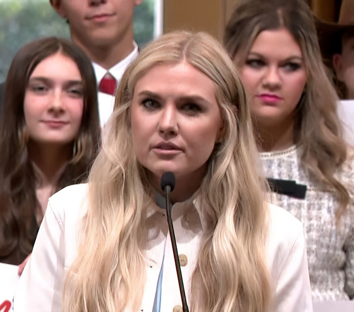 Blonde woman speaking at a microphone with a serious expression, highlighting Erika Kirk's latest video backlash.