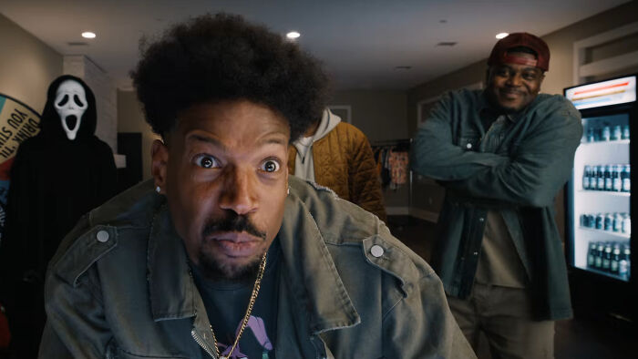 Man in a green jacket making a silly face with others in a room, scene from Scary Movie 6 trailer spoofing horror hit.
