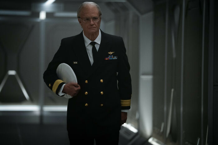 Actor in a naval uniform holding a white cap, portraying a character from For All Mankind season 5 in a dimly lit corridor.