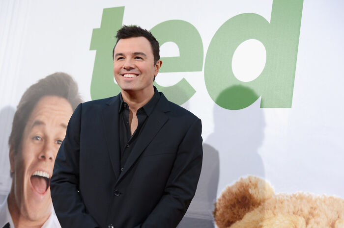 Seth Macfarlane at Ted event, smiling in black suit with Ted movie poster in background, hinting at Ted season 2 future.