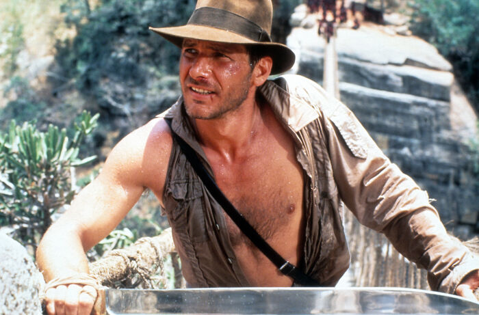 Harrison Ford as Indiana Jones, shirt partially open and wearing a hat, in an adventurous outdoor setting. Harrison Ford as Indiana Jones, shirt partially open and wearing a hat, in an adventurous outdoor setting.