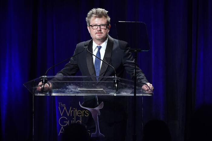 Man wearing glasses and a suit speaking at a podium during the Writers Guild Awards on a dark blue stage.