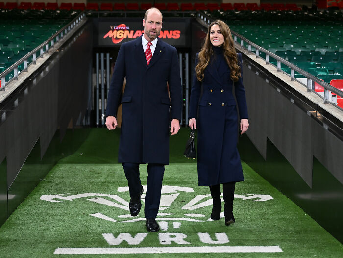 Prince William and Kate Middleton walking on a rugby field tunnel, attending a public event in formal coats.