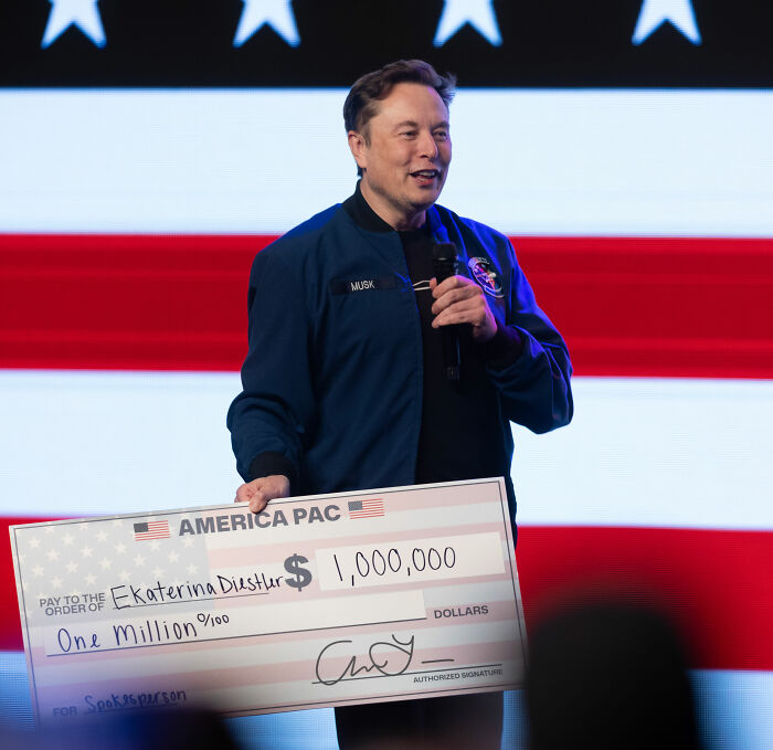 Elon Musk holding a large $1 million check on stage with an American flag background and speaking into a microphone